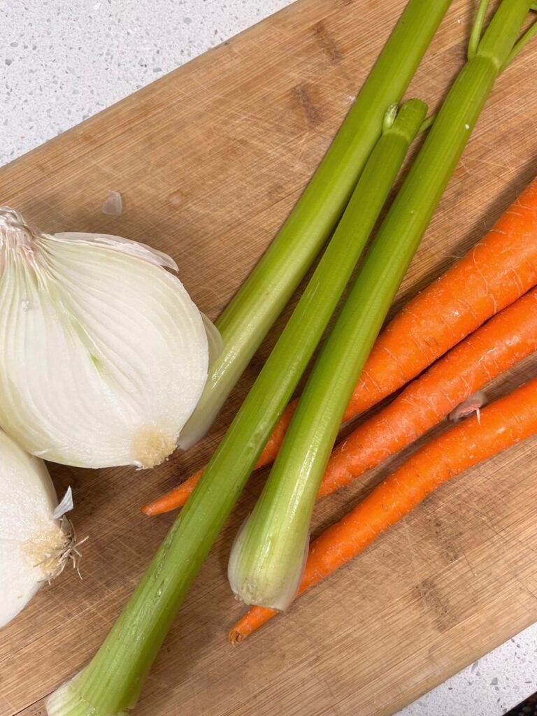 An onion cut in half, three ribs of celery, and three carrots on a wooden cutting board.