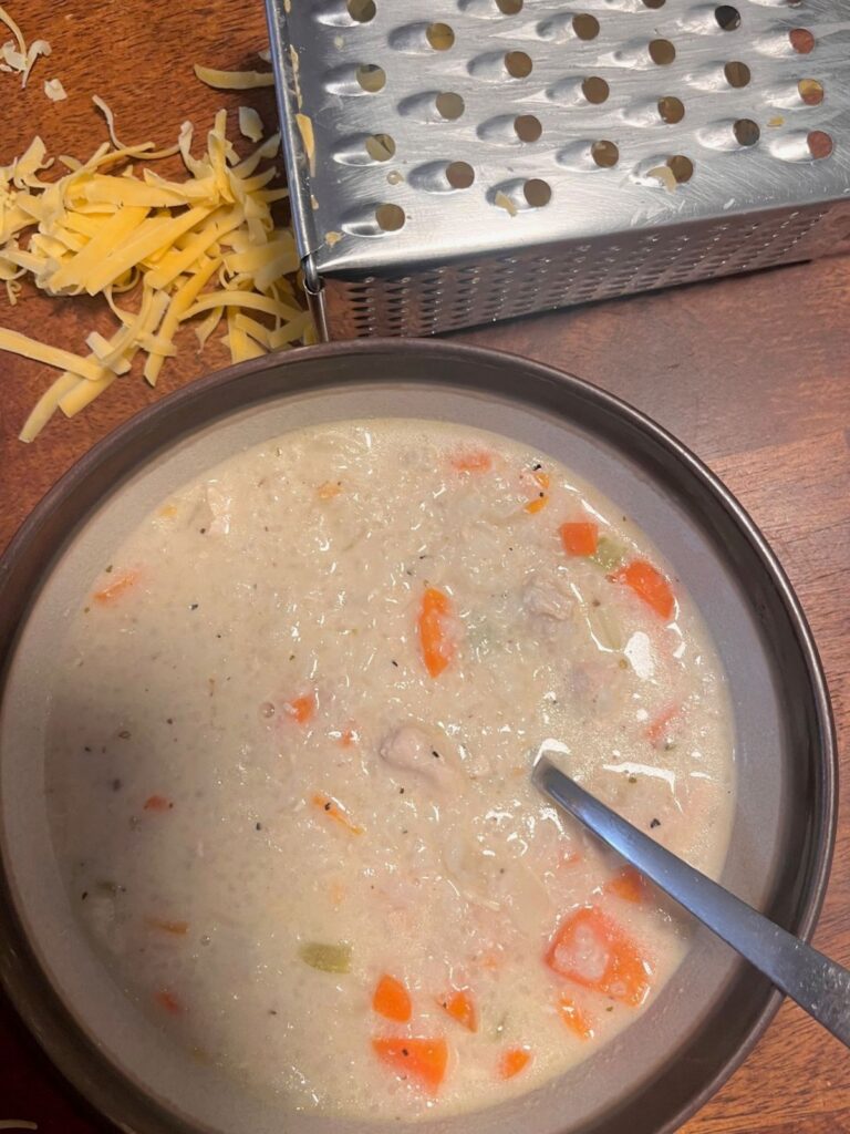 A bowl of soup next to a cheese grater and bits of cheddar.