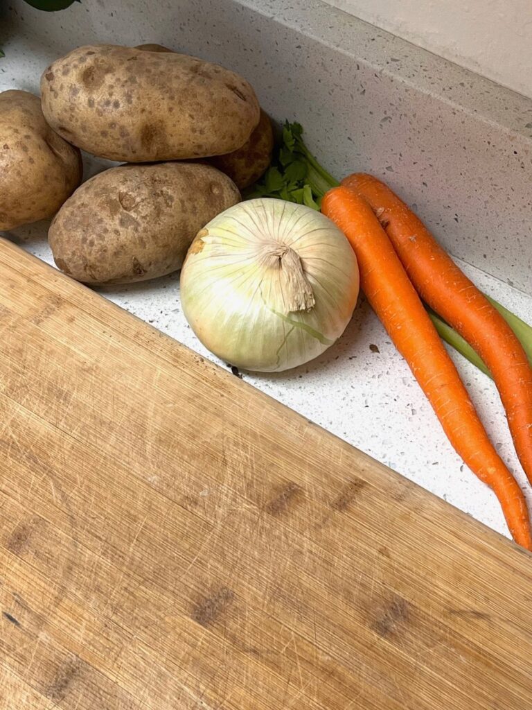 Carrots, potatoes, onions, and celery next to a worn cutting board. 