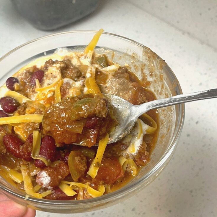 A glass bowl of homemade chili with a spoon, cheddar cheese, and sour cream.