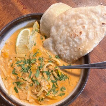 A bowl of chicken curry soup topped with cilantro, lime wedges, and a sourdough naan bread.