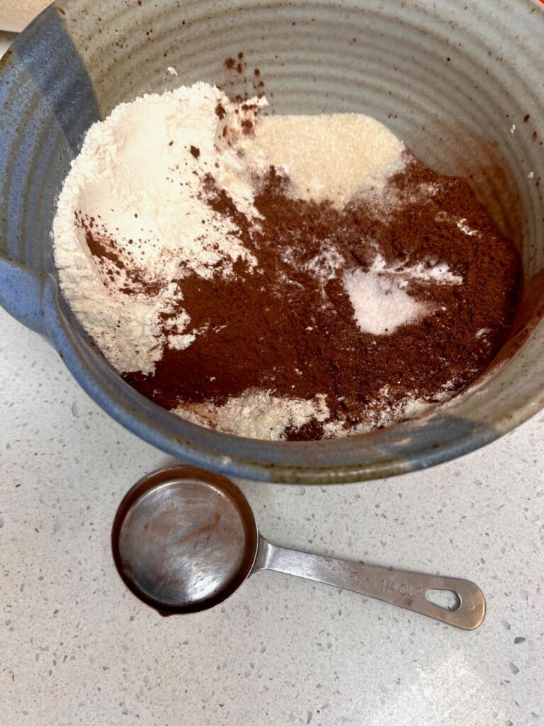 A bowl with dry ingredients and cocoa powder with a measuring cup next to the bowl.