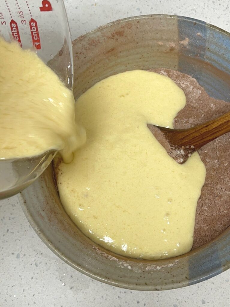 A glass liquid measuring cup pouring wet ingredients into a bowl of flour and cocoa powder with a wooden spoon. 