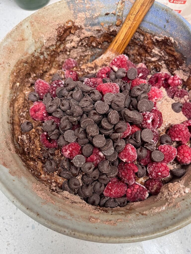 Chocolate sourdough muffin batter in a bowl with raspberries and chocolate chips poured on top with a wooden spoon. 