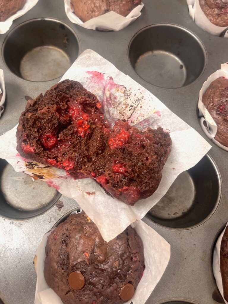 A chocolate muffin with bits of raspberry cut in half and laying open on a muffin tin.