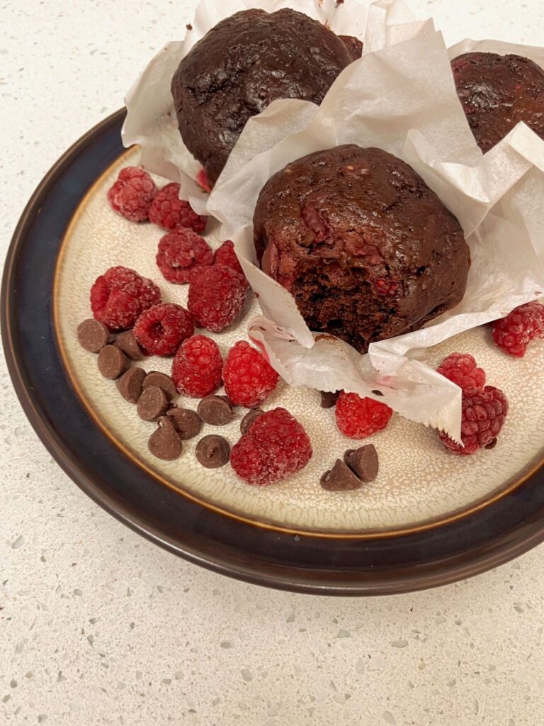 A dark muffin in a parchment paper liner sitting on a plate with red raspberries and chocolate chips.