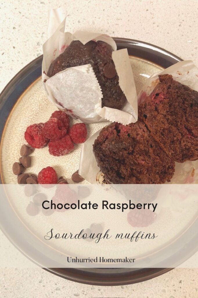 A chocolate raspberry sourdough muffin on a plate with another muffin still in the wrapper and raspberries and chocolate chips all on a plate. Unhurried homemaker.