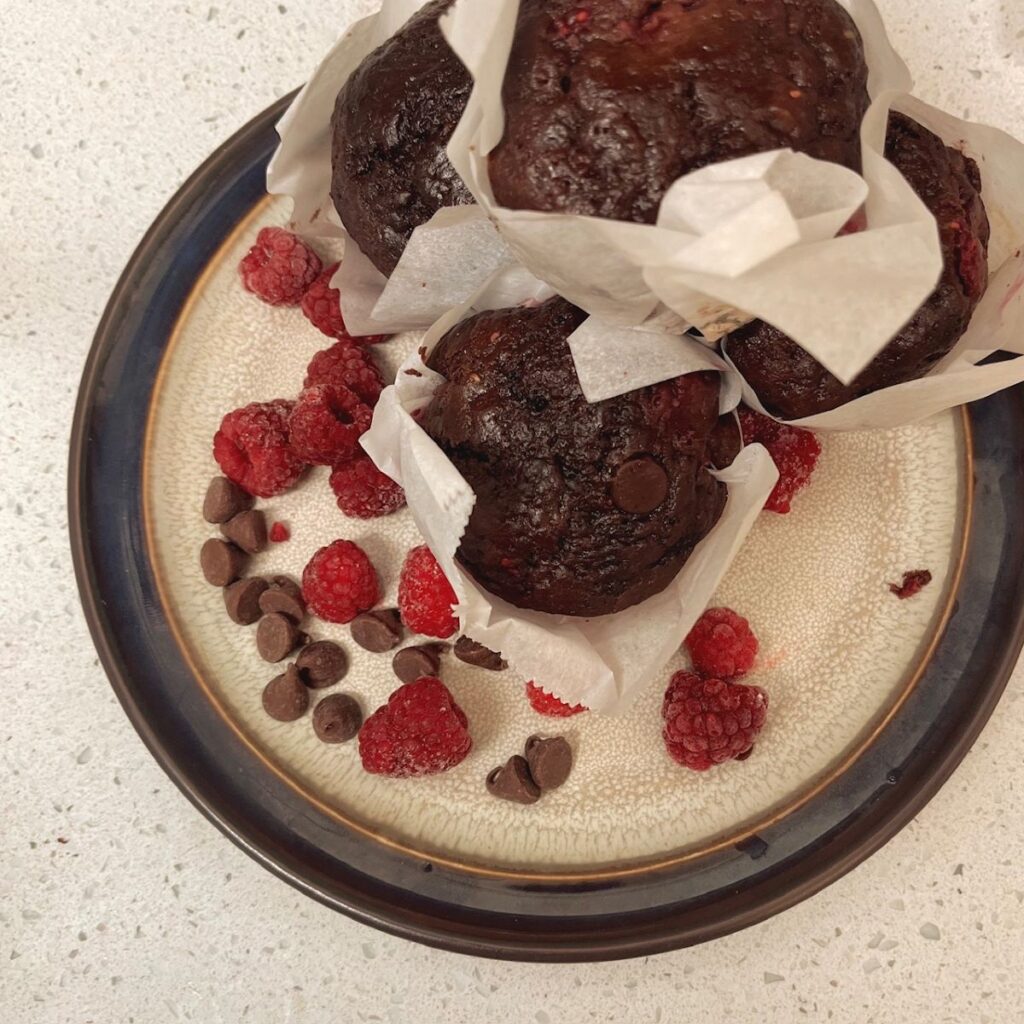 A pile of chocolate raspberry sourdough muffins on a plate with red raspberries and chocolate chips. 