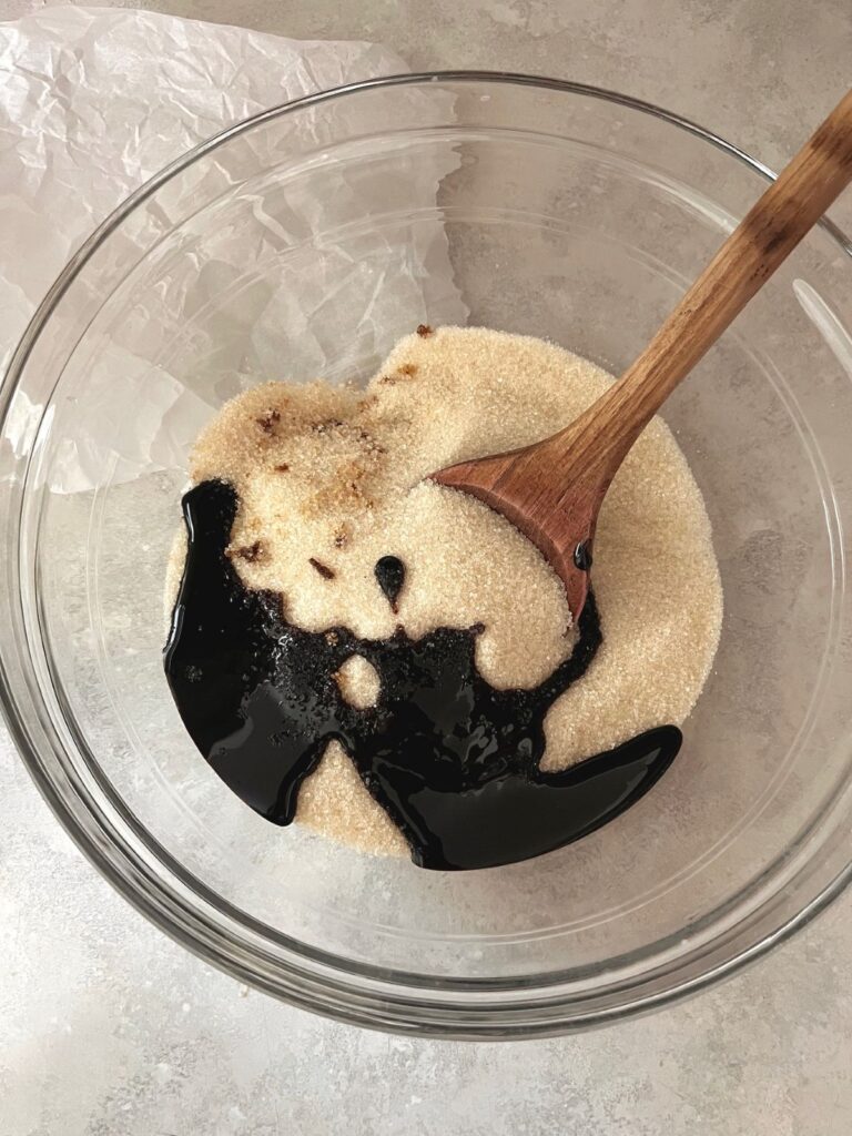 A glass bowl of white sugar with a drizzle of dark molasses and a wooden spoon.