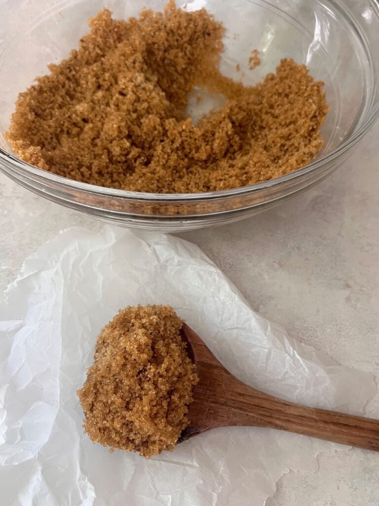 A bowl of brown sugar with a wooden spoon of sugar on a piece of parchment paper.